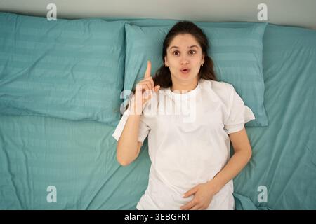 woman on bed with pillow pointing finger up with successful idea lying at home. number one. Stock Photo