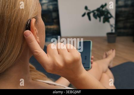 Young Hearing impaired woman using smartphone indoors. Close up of blonde woman wearing hearing aid on ear and pressing button while using smartphone Stock Photo