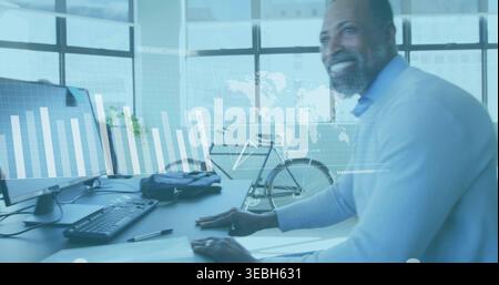 Smiling mature man wearing sweater analyzing charts on monitor with bicycle at office, copy space Stock Photo