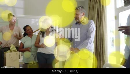 Gathering six adults chatting around dining table in living area, holding glasses and gift bag Stock Photo
