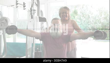 Doing lateral raises bearded man holding dumbbells in home gym, woman in pink top spotting Stock Photo