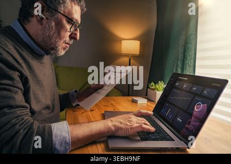 Senior man working at home desk analyzing financial report and digital dashboard on laptop. Warm indoor light, side view composition, focused expressi Stock Photo