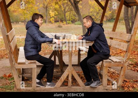 Wide shot of a man and his son sitting opposite each other in a wooden gazebo, playing chess surrounded by autumn leaves. Concept of quality time and Stock Photo