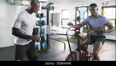 Pedaling man on air-resistance bike being coached at gym, wearing gray tee and black shorts Stock Photo