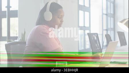 Working woman wearing white headphones and pink shirt focusing on laptop at office desk, banding Stock Photo