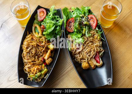 Asian noodle dishes with salad and beer. Top view of two Asian noodle plates with fresh salad, tomatoes, vegetables, seafood, and beer on a wooden Stock Photo
