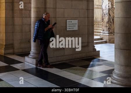 A senior man checks his phone leaning against a stone column in an elegant marble-floored corridor with directional signage, Victoria & Albert Museum Stock Photo