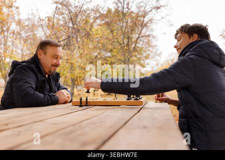 Close up of a smiling teen boy making a chess move during a game with his father in an autumn park. Focus on the hand and decorative chess pieces. Str Stock Photo