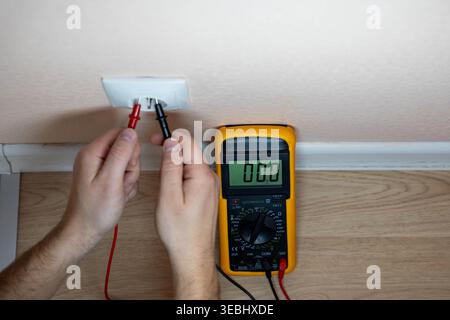 Hands Testing Wall Outlet With Multimeter, DIY Homeowner Performing Safety Check Near Baseboard, Probes Inserted Into Faceplate, Digital Meter Reading Stock Photo