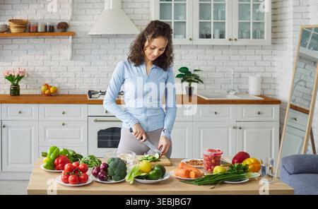 Fit sporty woman in modern home kitchen, chopping vegetables on cutting board, fresh colorful meal  Stock Photo