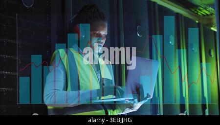 Working Black technician in hi-vis vest typing on laptop in server aisle with headset and overlays Stock Photo
