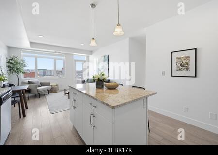 Modern kitchen with white cabinets and granite countertop. Open floor plan flows to bright living room. Large windows bring in natural daylight. Light Stock Photo