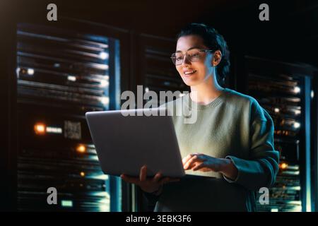 A professional woman in glasses stands confidently with a laptop in a high-tech data center, illuminated by the glow of server racks. This image conve Stock Photo