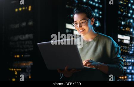 Confident young woman in glasses works on a laptop in a high-tech server room. Surrounded by glowing server racks, she exudes happiness and profession Stock Photo