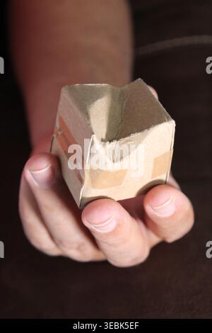 A man's hand holding a damaged miniature cardboard box with brown tape; at the sofa's arm. Concept: A client received a damaged package at home Stock Photo