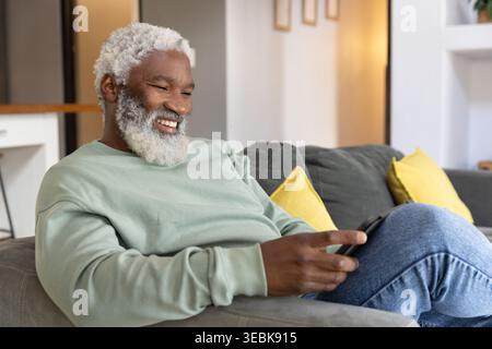 Senior man sitting on gray sofa in living room holding phone, wearing sweatshirt, copy space Stock Photo