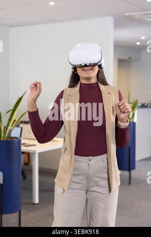 Mid adult woman standing interacting with white VR headset in office wearing maroon top, beige vest Stock Photo