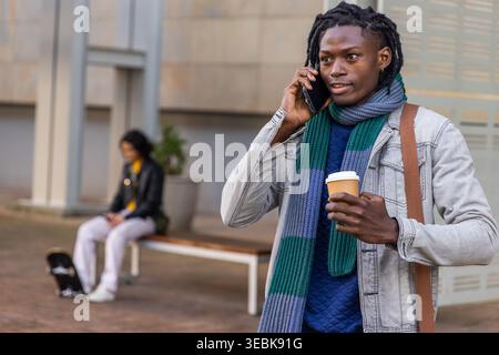 African American man wearing denim jacket, scarf, talking on smartphone holding takeaway coffee cup Stock Photo