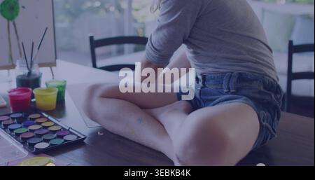Sitting girl painting on craft table at home, in gray top denim shorts with paint pots Stock Photo