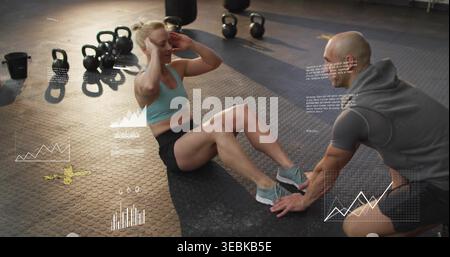 Doing crunches, trainee in sports bra on rubber floor, trainer bracing feet, kettlebells, metrics Stock Photo