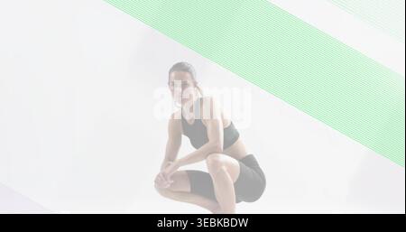 Posing athlete in low squat in studio, wearing black sports bra, shorts, hoops, mint-lavender bands Stock Photo