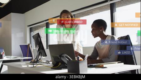 Collaborating two women leaning over laptop at office desk, white blouse, jacket, notifications Stock Photo