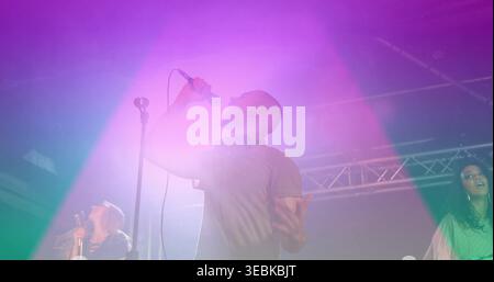 Singing bald male singer holding mic with headphones on concert stage, magenta purple green beams Stock Photo
