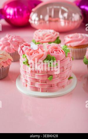 A pink cake decorated with a basket of roses and delicate butterfly embellishments, surrounded by pink cupcakes Stock Photo