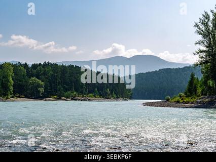Light blue river flows through a dense forest with layered mountains visible in the distance on a bright summer day Stock Photo