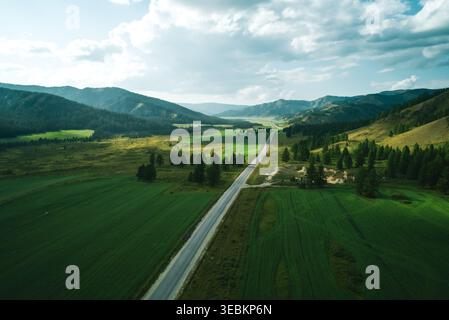 Long straight road cutting through a vast green valley with forested mountains under a cloudy sky Stock Photo