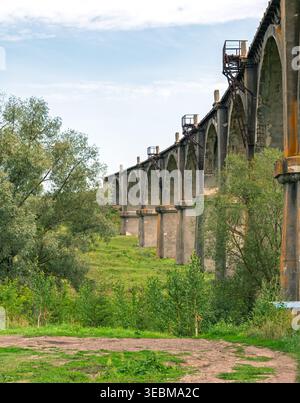 Abandoned concrete railway viaduct with arches standing on green grassy hills Stock Photo