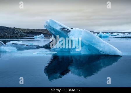 View of a luminous blue iceberg drifting serenely in the glacial lagoon, its jagged edges reflecting in the calm waters, Glacier Lagoon, Sveitarfelagi Stock Photo