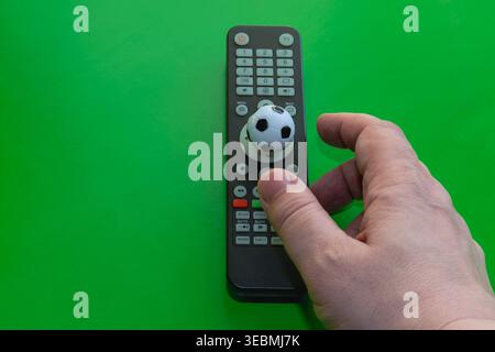 Male hand reaching for remote with small soccer ball on bright green broadcast concept. Stock Photo
