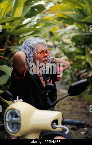 Mature woman on scooter adjusting hair on tropical road, Canggu, Bali Stock Photo