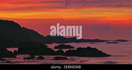 Fiery sunset sky over dark rocky coastline silhouettes and calm water Stock Photo