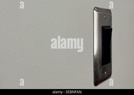 A light switch is mounted on a plain wall. The switch is in the off position. It is a common type used in many buildings. The room has natural light e Stock Photo