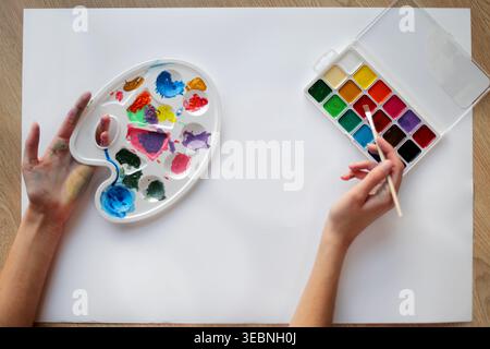 Hands Painting With Colorful Art Supplies. Overhead view of hands using paintbrush, palette, and watercolor set on blank paper for a creative art Stock Photo