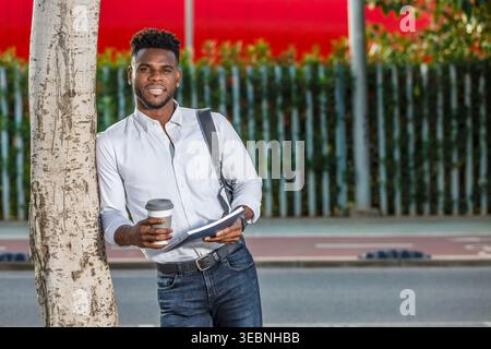 An African American student stands next to a tree, cup of coffee in hand. Stock Photo