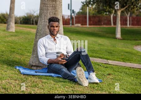 An African American man sits quietly under a tree while using his cell phone and cup of coffee. Stock Photo