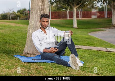 A man sits comfortably in the green grass, leaning against a tall tree, enjoying the warmth of a sunny day while holding a cell phone and a cup of cof Stock Photo
