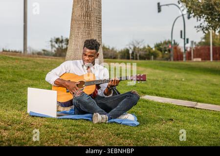 A man in casual clothes is sitting on the green grass and playing a guitar. Stock Photo