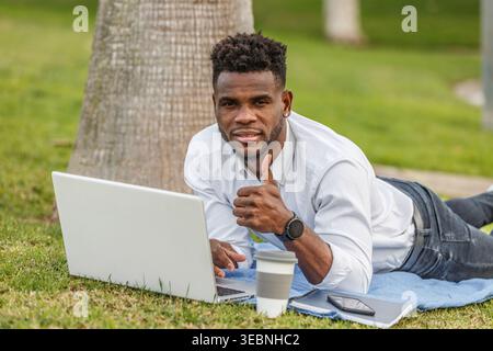 An African-American man is seen with his thumb up lying on the floor while using a laptop to work. Stock Photo