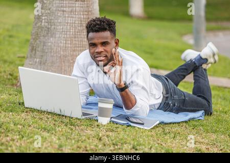 A man doing the okey symbol with his hand seen on the ground using a laptop to work, absorbed in his tasks. Stock Photo
