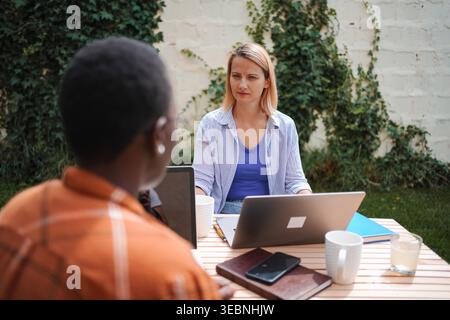 Two businesswomen working remotely outdoors, using laptops and discussing a project Stock Photo