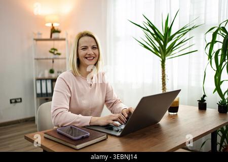 A cheerful professional engages with her laptop in a sunlit office, surrounded by greenery and personal touches. Warm vibes fill the space as she enjoys her task. Stock Photo