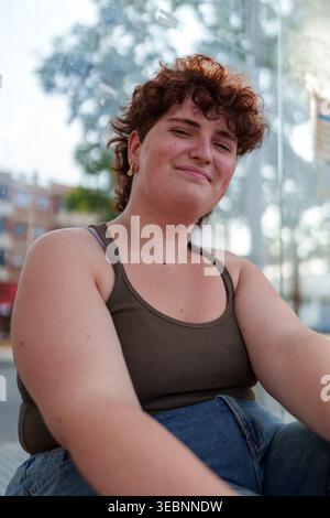 Queer young adult looking at camera, feeling positive and authentic while sitting in a modern city environment Stock Photo