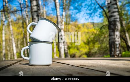 Two white enamel camping mugs stacked on wooden table in spring birch forest Stock Photo