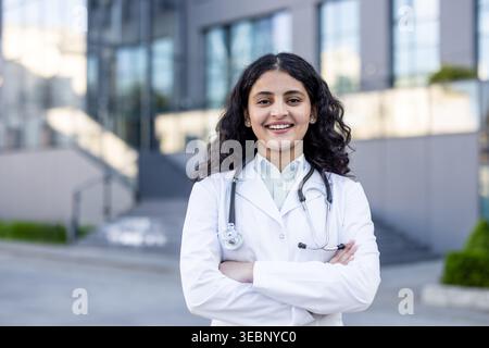 Young indian female doctor in a white lab coat standing with arms crossed, smiling confidentially in front of a modern hospital building, representing healthcare, professionalism, and medical career Stock Photo