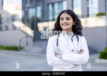 Young indian female doctor wearing a white lab coat and stethoscope, smiling confidently with arms crossed while standing outside a modern hospital building Stock Photo
