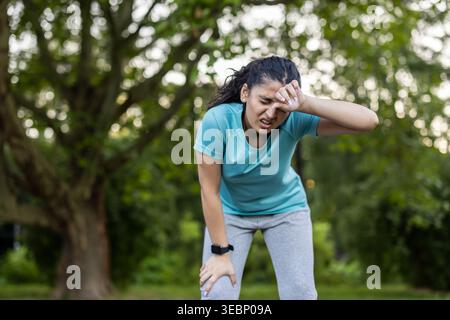 Exhausted young woman standing outdoors, wiping sweat from her forehead after an intense fitness workout, experiencing fatigue and shortness of breath during her exercise routine Stock Photo